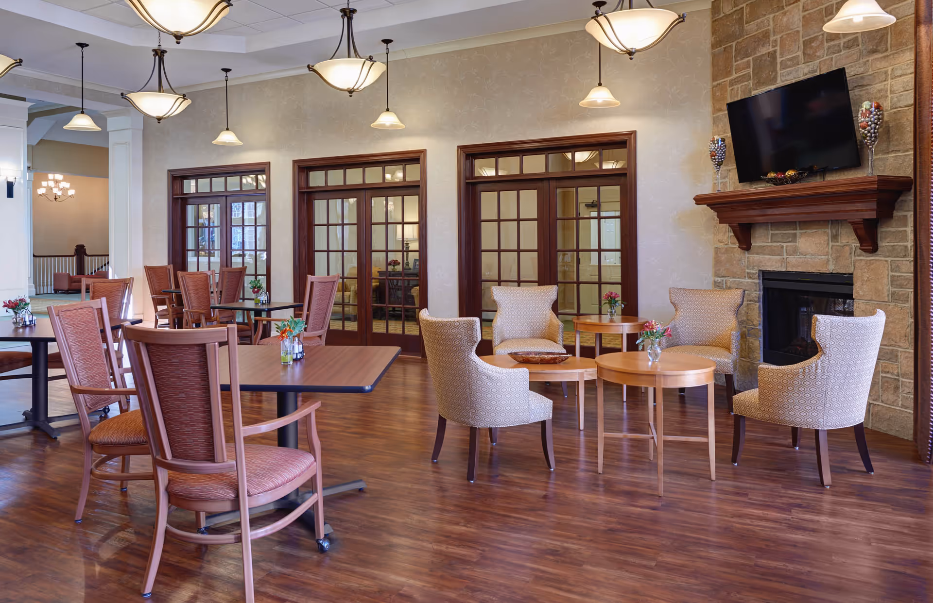 A spacious and well-lit common area with wooden flooring featuring several tables and chairs arranged for dining and socializing. There are upholstered armchairs around small round tables near a stone fireplace with a mounted flat-screen TV above it. The room has multiple hanging light fixtures and large wooden-framed glass doors leading to another room.