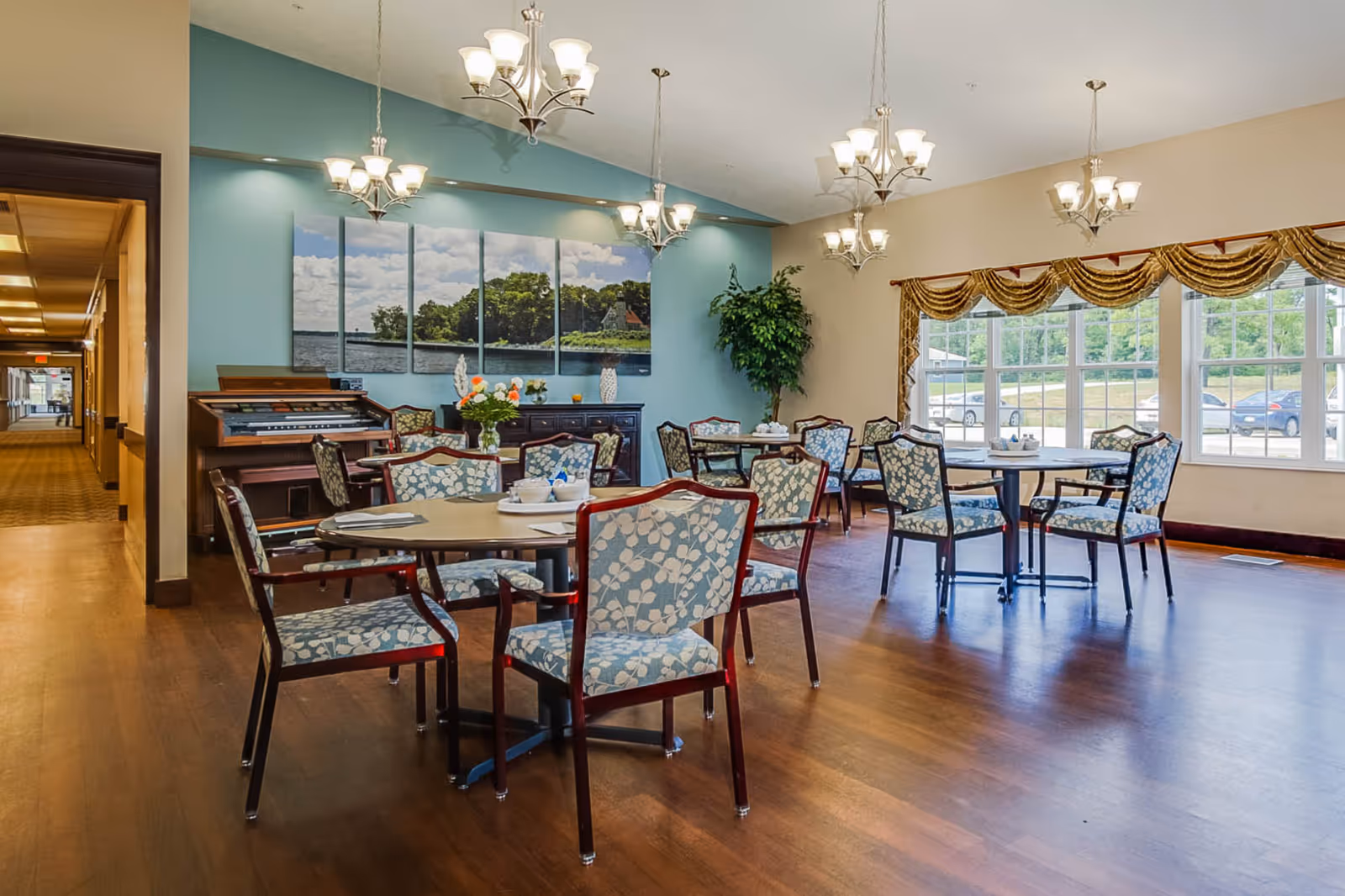 A dining room in an assisted living facility with several round tables surrounded by chairs upholstered in a blue and white floral pattern. The room features wooden flooring, large windows with gold valances, and multiple chandeliers hanging from the ceiling. A blue accent wall displays a multi-panel artwork of a lakeside scene, and there is a piano and a plant in the corner.