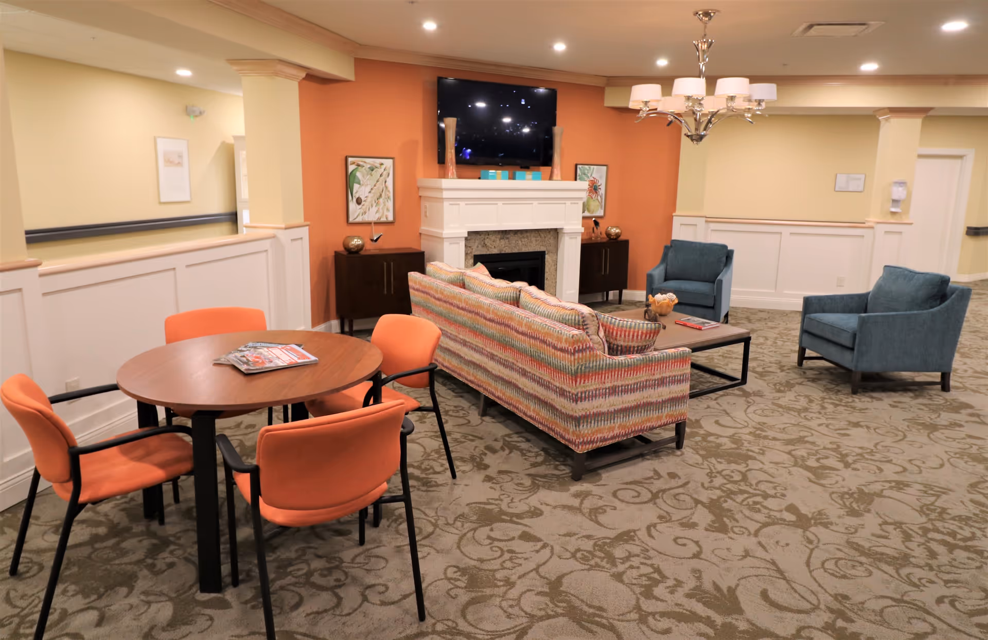 A cozy senior living facility common area with a patterned sofa, two blue armchairs, a round wooden table with four orange chairs, a fireplace with a mounted TV above it, and decorative artwork on the walls. The room has warm lighting and a patterned carpet.