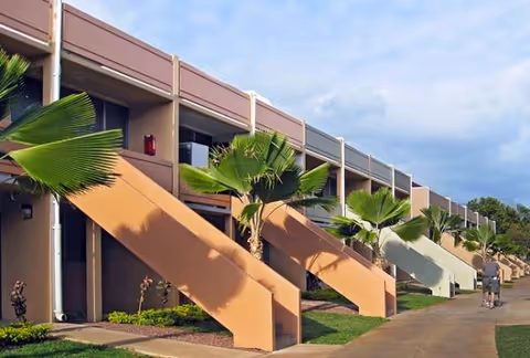 Exterior view of Kukui Gardens building with ramps, balconies, palm trees, and a pedestrian.