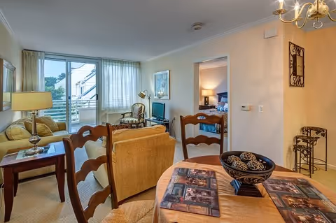 A cozy living and dining area in a senior living facility with a wooden dining table and chairs in the foreground, a beige sofa and armchair near a window with sheer curtains, a small TV on a stand, and a view into a bedroom with a bed and nightstand lamp.