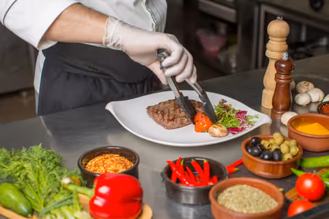 A chef wearing gloves is plating a cooked steak with grilled vegetables and salad on a white plate in a kitchen setting. Various fresh ingredients including red chili peppers, olives, tomatoes, and spices are arranged on the countertop nearby.