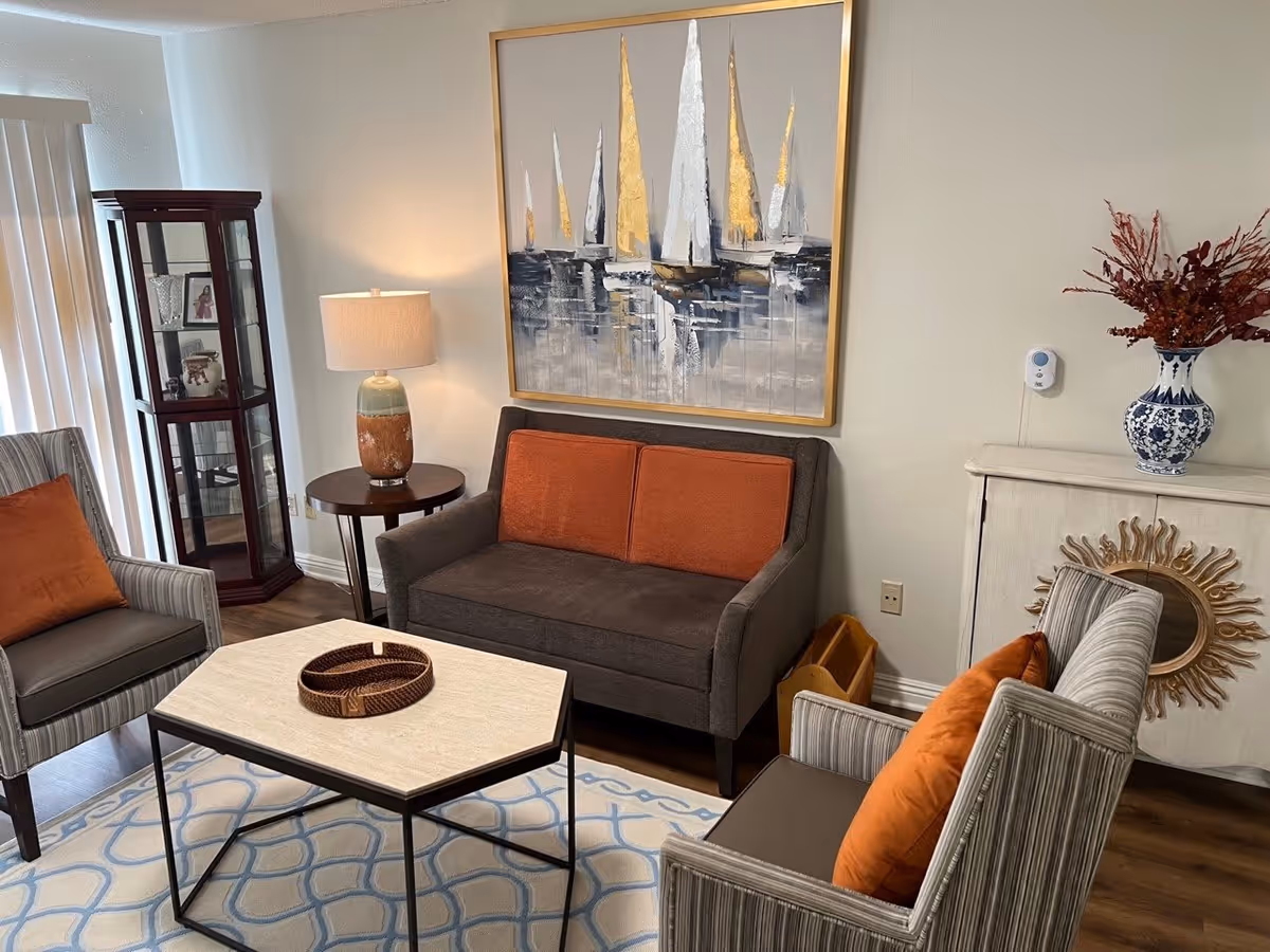 A cozy living room area with a small dark gray loveseat with orange cushions, two striped armchairs with orange pillows, a light-colored coffee table with a decorative tray, a round side table with a lamp, a glass display cabinet, a white cabinet with a sunburst mirror decoration, and a large painting of sailboats on the wall.