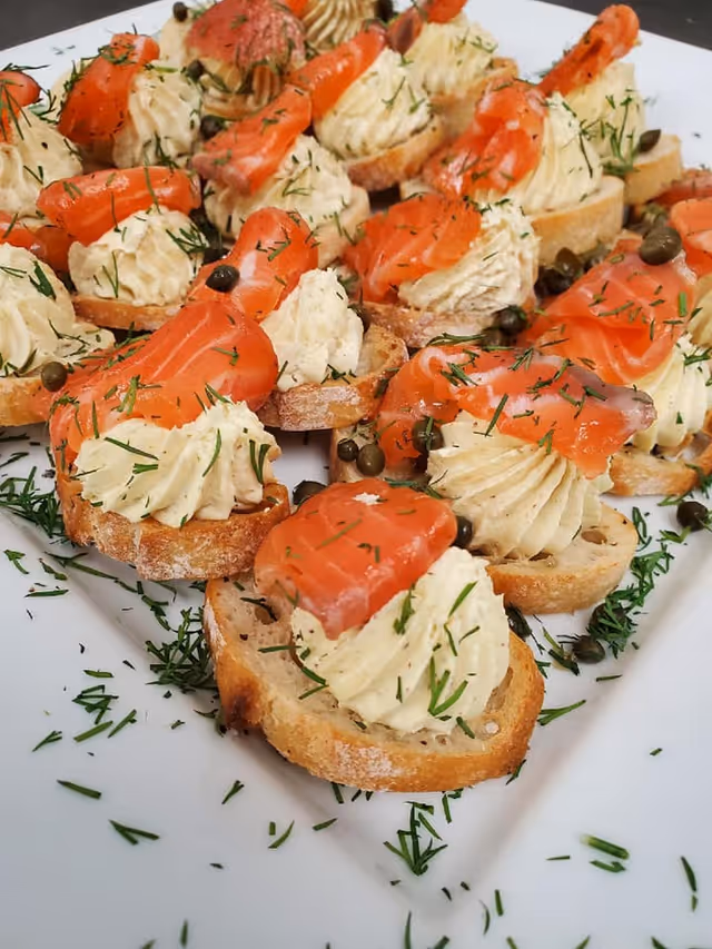 Close-up of small toasted bread slices topped with a swirl of creamy spread, smoked salmon, capers, and sprinkled with fresh dill on a white plate.