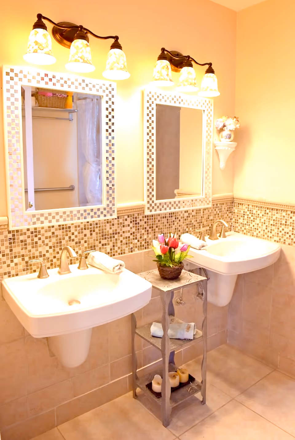 A bathroom with two white pedestal sinks side by side, each with a towel placed on the edge. Above each sink is a rectangular mirror with a mosaic tile border and a light fixture with three lamps. Between the sinks is a small metal stand holding a basket of colorful tulips, rolled towels, and candles. The walls are tiled halfway up with small square mosaic tiles and painted a light beige color above.