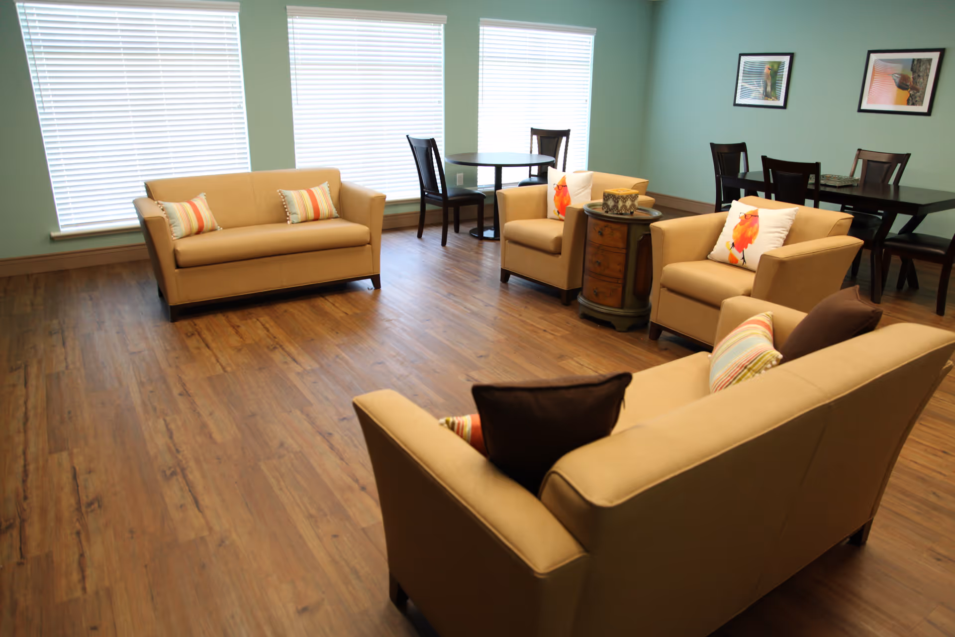 Bright communal living room with beige sofas and armchairs arranged on wood floors, small dining tables, and large windows with blinds.