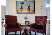 Two red patterned chairs flank a small table with a vase and wall art.