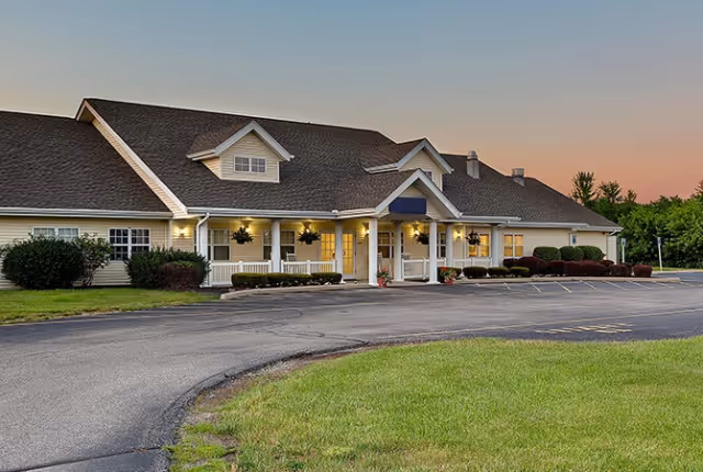 Exterior view of a single-story senior living facility building with a large covered entrance, well-maintained landscaping, and a paved driveway at sunset.