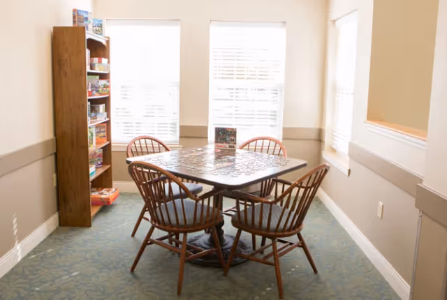 Small activity room with a square table and four wooden chairs around a partially completed jigsaw puzzle, a shelf of games, and three bright windows.