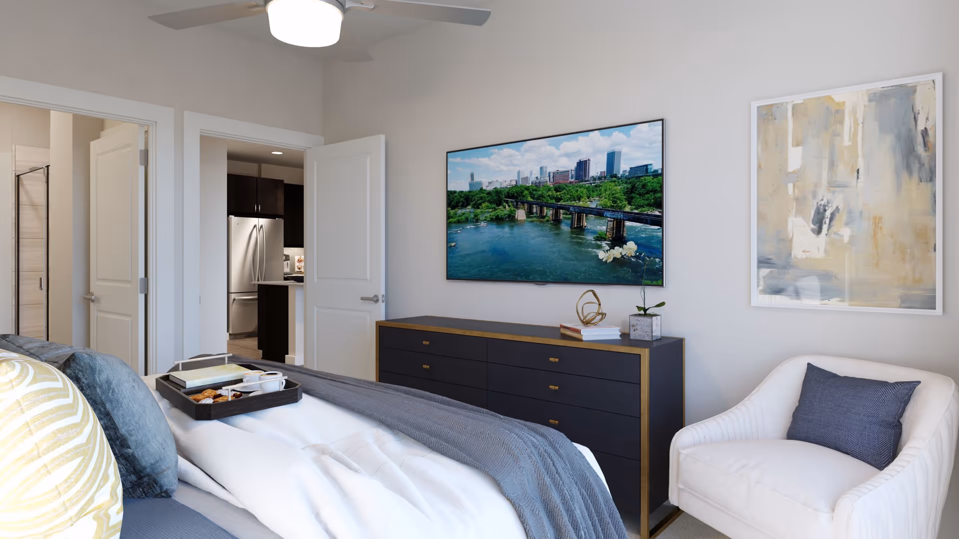 A modern bedroom with a bed featuring multiple pillows and a tray with breakfast items. There is a white armchair with a dark cushion, a black dresser with decorative items, and a large flat-screen TV mounted on the wall displaying a scenic river and city skyline. Two open doors reveal a kitchen area with a stainless steel refrigerator and a bathroom.