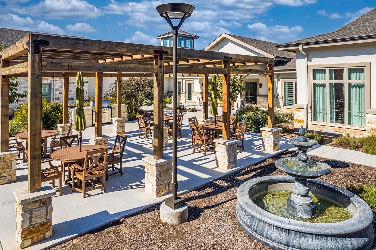 Outdoor patio area at Sage Valley Senior Living featuring wooden pergolas with tables and chairs underneath, green umbrellas, a stone water fountain in the foreground, and a building with large windows in the background under a blue sky with scattered clouds.