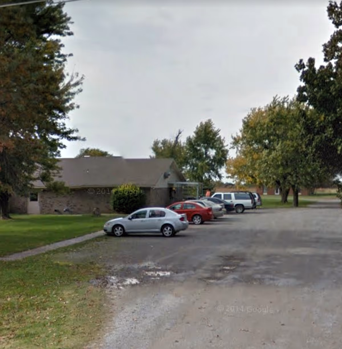 Parking lot with several parked cars in front of a single-story brick building surrounded by trees.