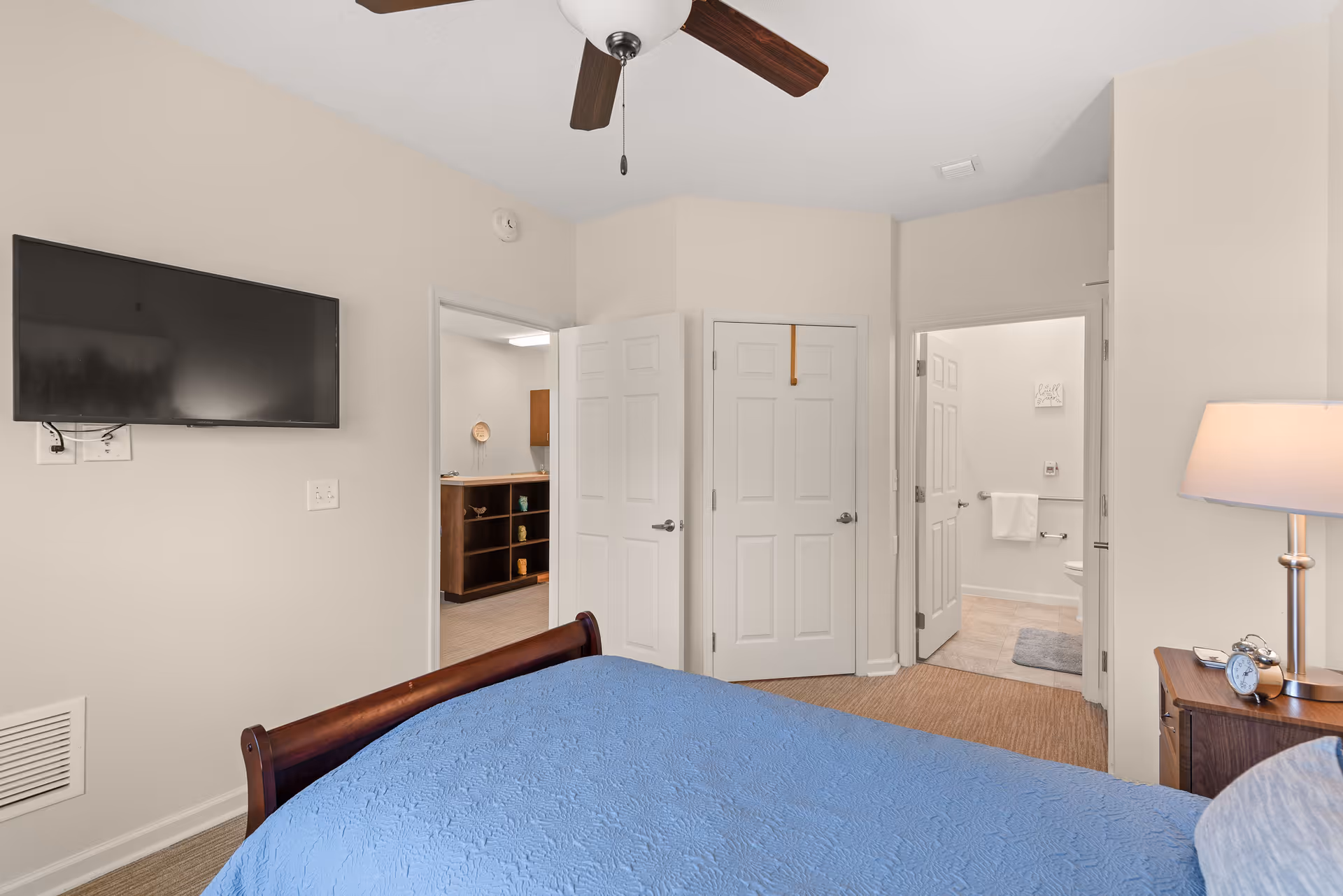 A bedroom with a wooden bed frame covered with a blue quilt. On the right side, there is a wooden nightstand with a lamp and an alarm clock. A ceiling fan with dark wooden blades is mounted on the ceiling. A flat-screen TV is mounted on the wall to the left. There are two open doors, one leading to a bathroom with a towel rack and toilet visible, and the other leading to another room with a wooden shelf unit.