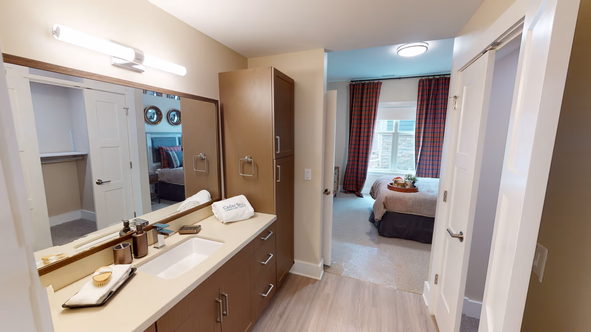 View of a senior living facility bathroom with a large mirror, countertop with sink, and wooden cabinets. The bathroom opens into a bedroom with a bed, plaid curtains, and a window letting in natural light.