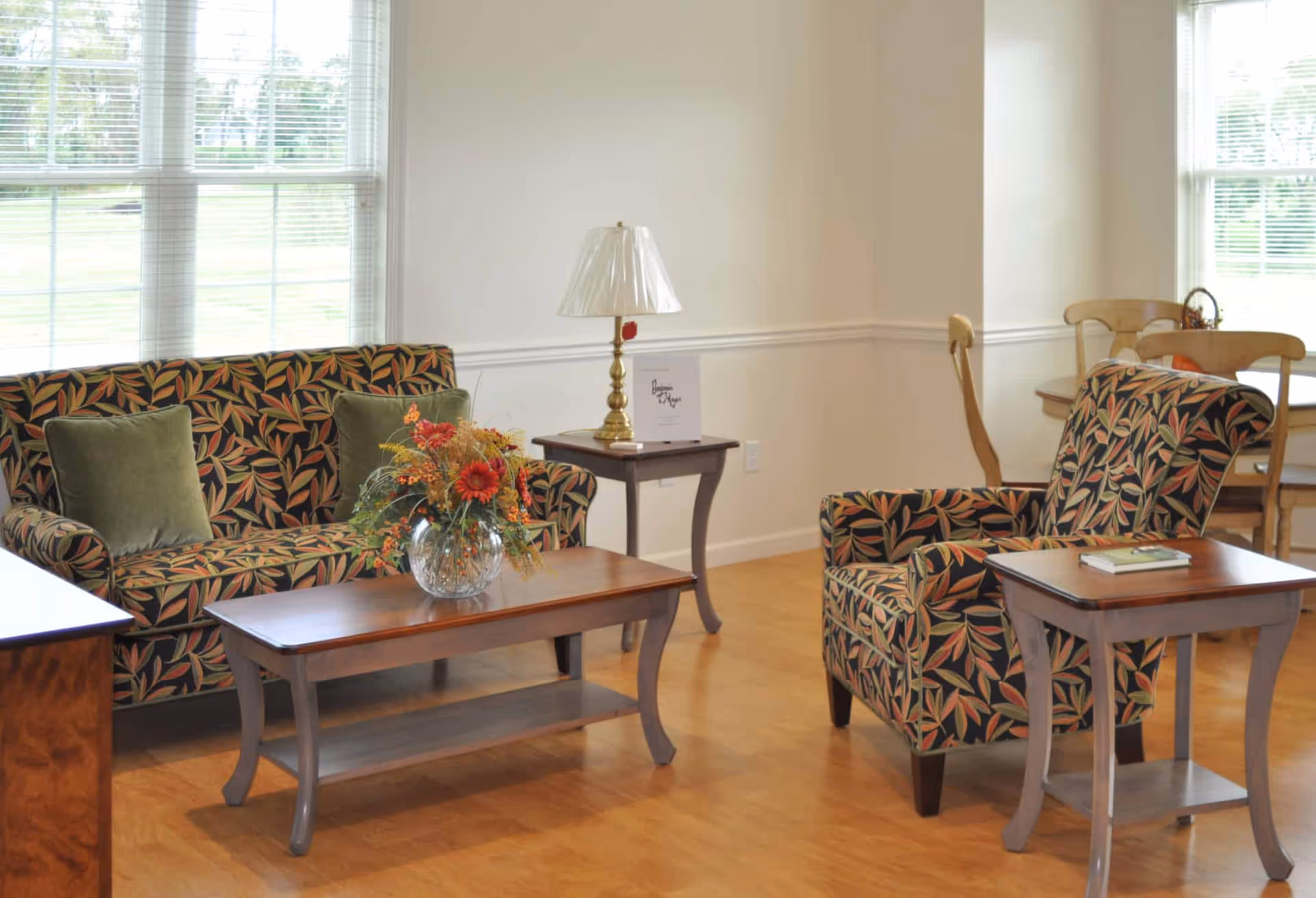 Bright living room with a patterned sofa and armchair, wooden coffee and side tables, a lamp, and a floral centerpiece by large windows.