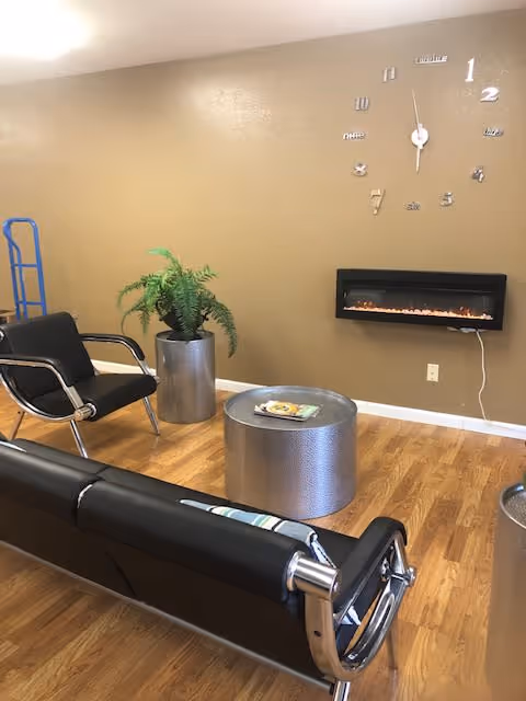 Waiting room/lounge with black modern chairs, a silver coffee table and planter, a wall-mounted electric fireplace and a decorative clock on a tan wall.