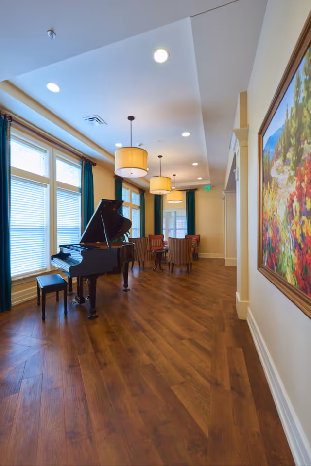 A bright and spacious room with large windows covered by teal curtains, featuring a black grand piano with a matching bench on the left side. The room has wooden flooring and several pendant lights hanging from the ceiling. In the background, there are several striped upholstered chairs arranged around small tables. A colorful landscape painting is visible on the right wall.