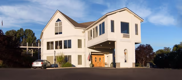 Exterior view of a two-story white building with large windows and a covered entrance. There is a car parked on the left side and trees surrounding the building under a clear blue sky.