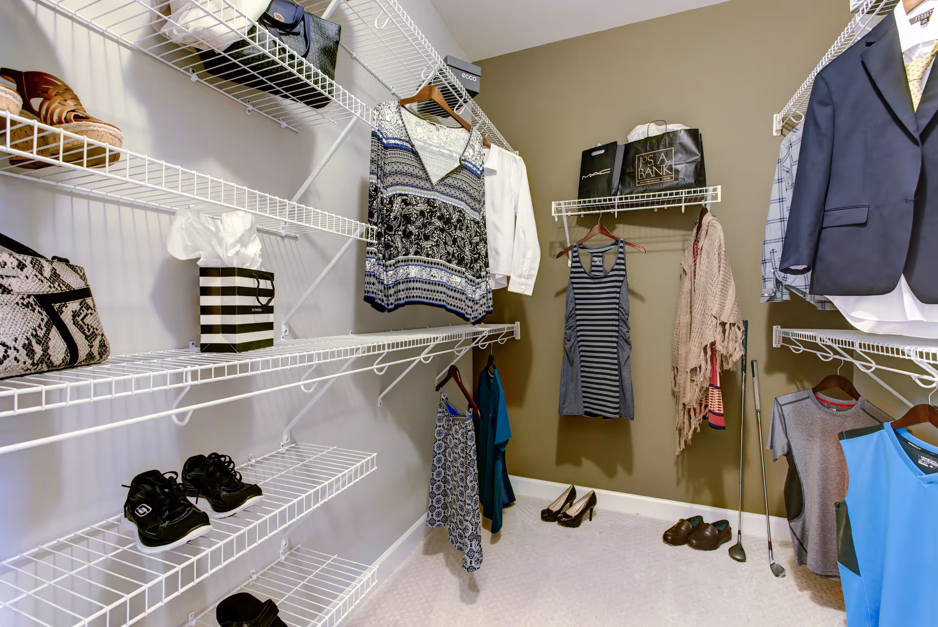 A walk-in closet with white wire shelving on three walls. Various clothing items including dresses, shirts, jackets, and shoes are neatly arranged on hangers and shelves. The walls are painted beige, and the floor is carpeted in a light color.