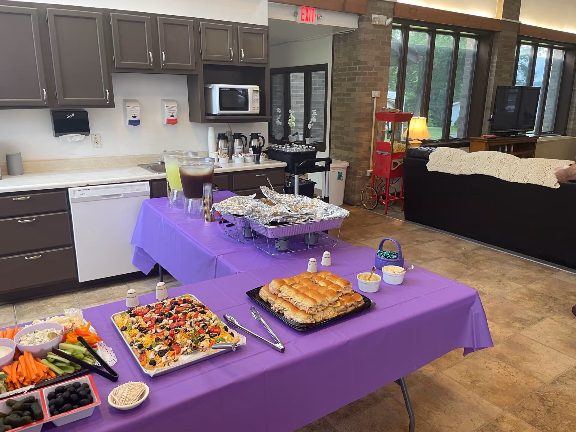 A kitchen and living room area in a senior living facility with a purple tablecloth-covered table set with trays of sandwiches, a vegetable platter with dip, and a tray of nachos topped with olives and tomatoes. Behind the table, there are pitchers of lemonade and iced tea, a microwave, coffee pots, and a popcorn machine near large windows letting in natural light. A black couch with a white knitted throw is partially visible in the living room area.