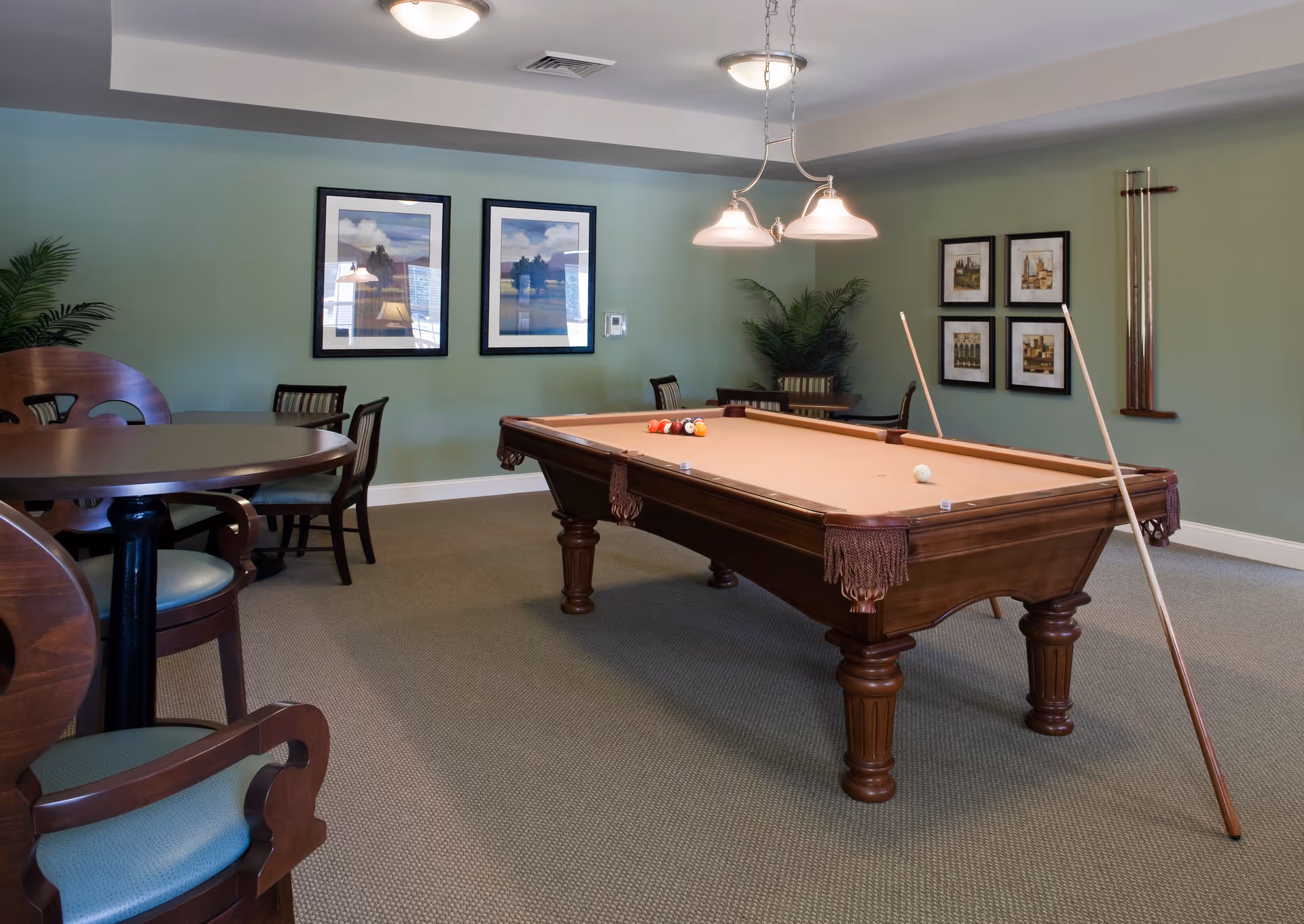 A recreational room with a pool table in the center, pool cues resting on the table, and a set of billiard balls arranged on the felt surface. The room has green walls adorned with framed artwork and a rack holding additional pool cues. There are several wooden chairs with blue cushions and round tables around the room. Ceiling lights illuminate the space.