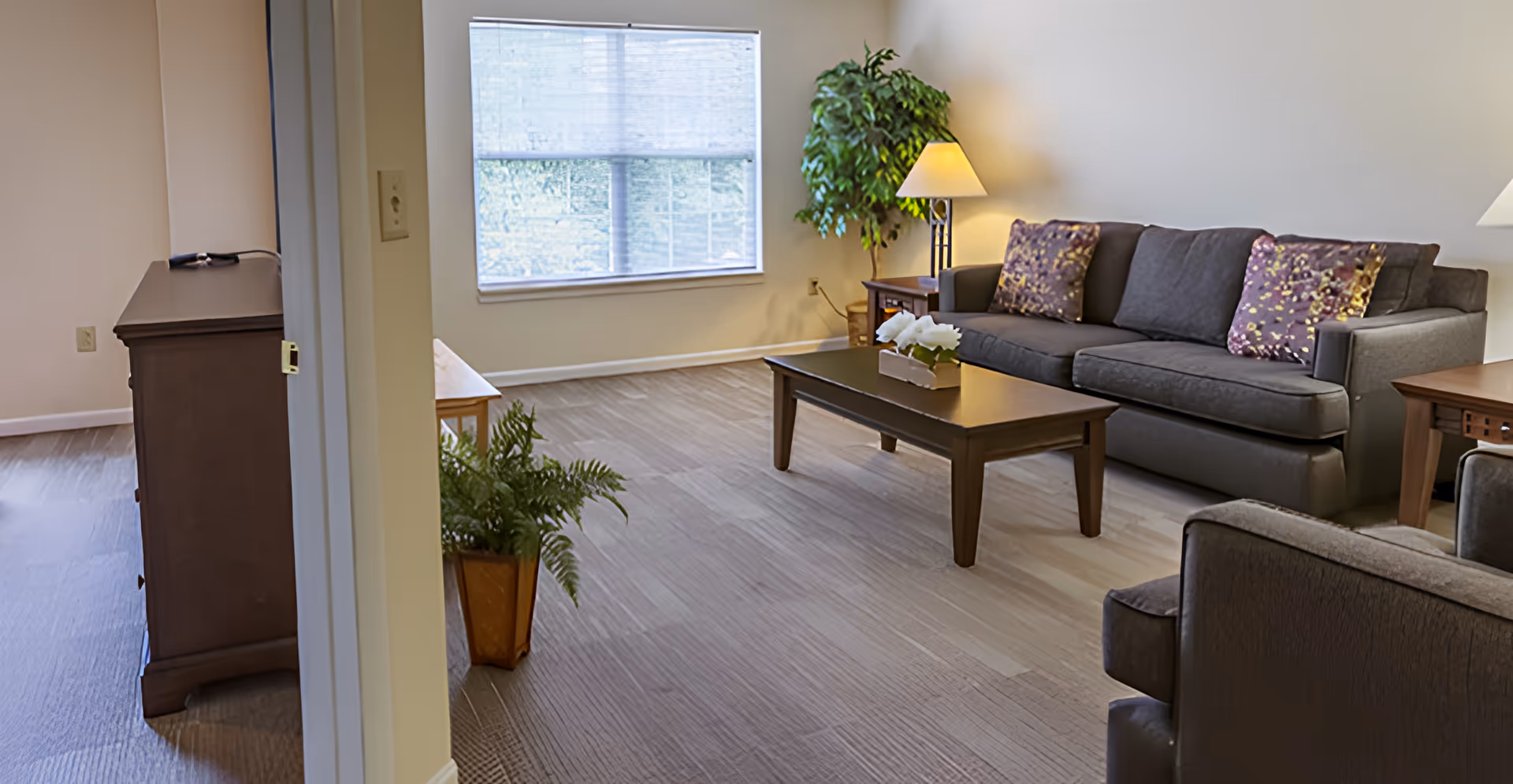 Bright furnished living room with a sofa, coffee table, armchair, lamps, and plants near a large window.