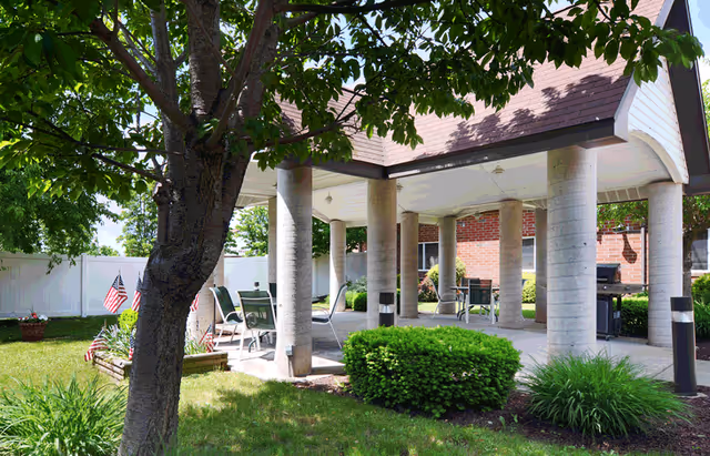 Outdoor covered patio area with several chairs and tables surrounded by green bushes and trees, with a brick building and white fence in the background. American flags are placed in the garden beds.