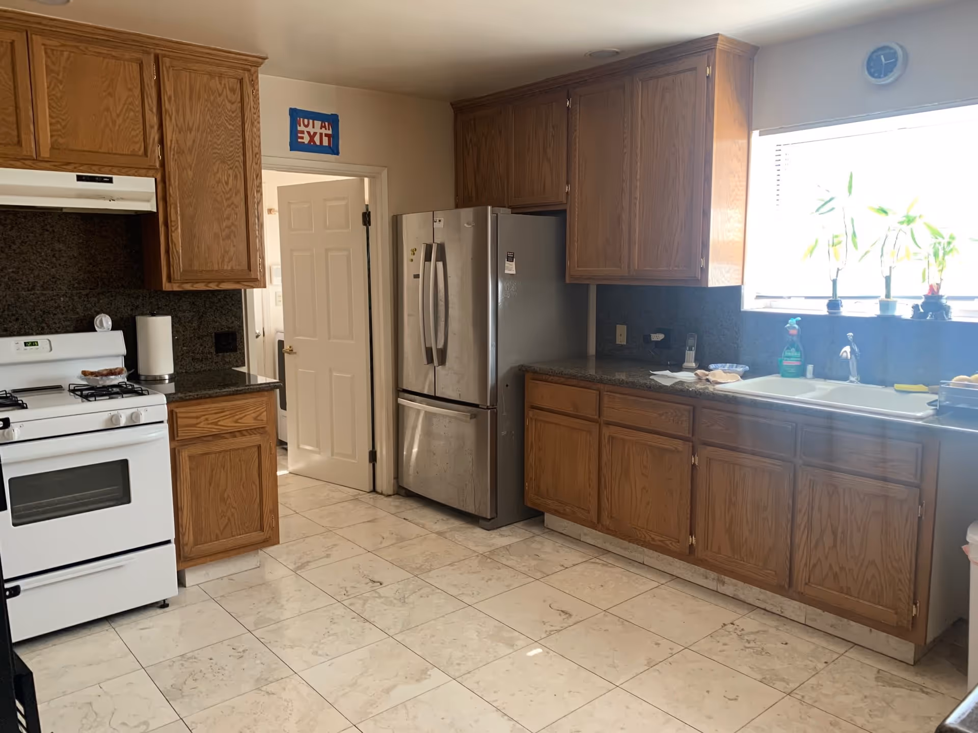 A kitchen with wooden cabinets, a white gas stove, a stainless steel refrigerator, a double sink under a window with potted plants, and a tiled floor. There is a door slightly open with a sign above it that reads 'NOT AN EXIT'.