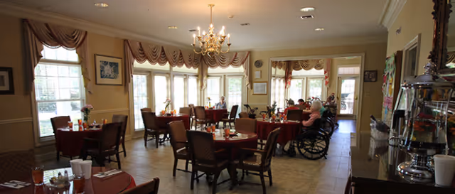 A spacious dining room with multiple round tables covered with red tablecloths, each set with glasses, plates, and condiments. Several chairs surround the tables, and a few people are seated, including one person in a wheelchair. Large windows with draped curtains allow natural light to fill the room. A chandelier hangs from the ceiling, and there is a serving area with a beverage dispenser on the right side.