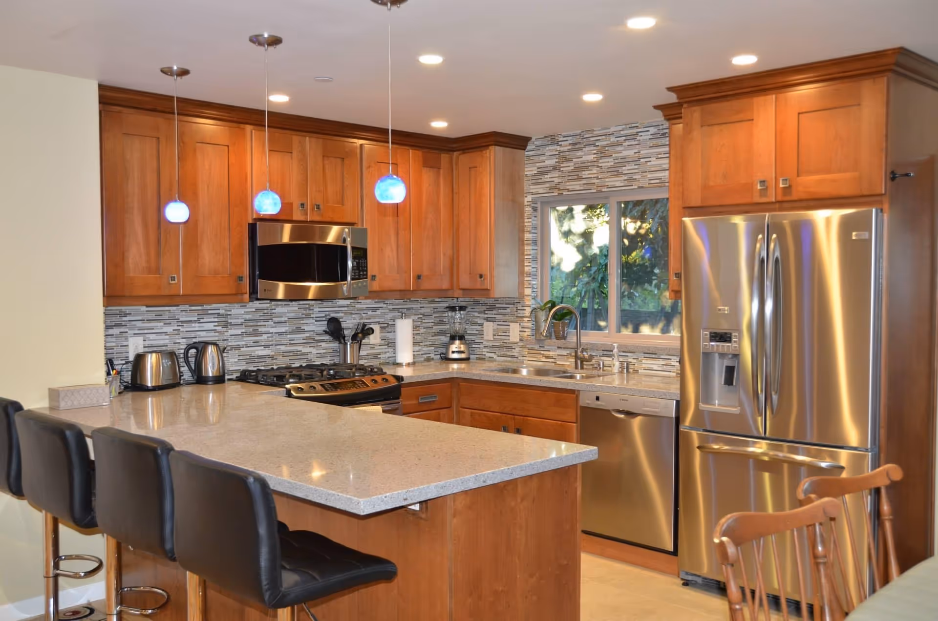 Modern kitchen with wooden cabinets, a large island with four black cushioned bar stools, stainless steel appliances including a refrigerator, microwave, dishwasher, and stove. The backsplash features a mosaic tile design, and three blue pendant lights hang above the island. A window above the sink shows greenery outside.