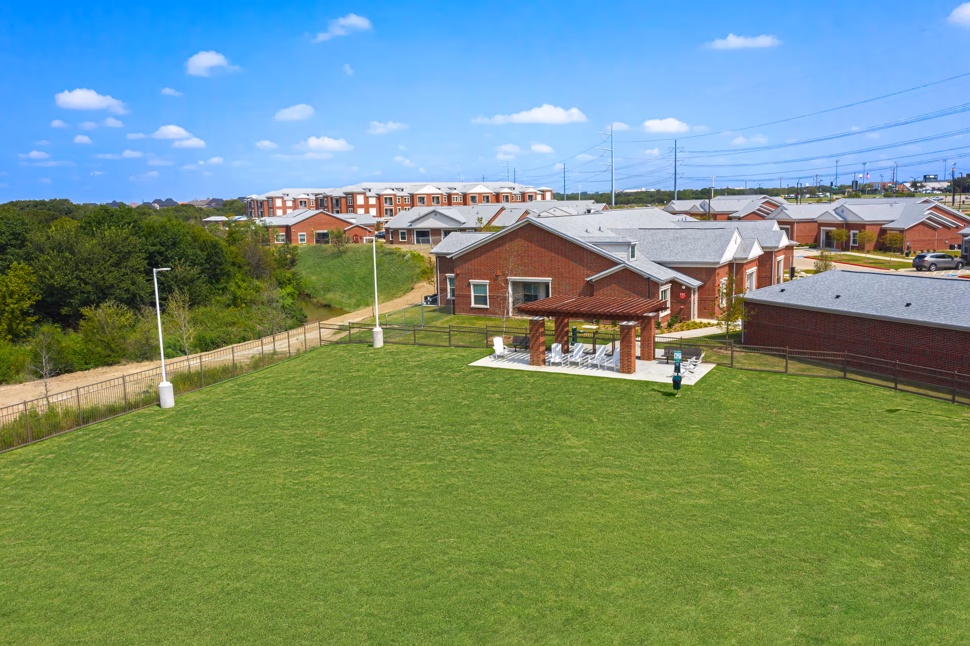 Aerial view of a senior living facility showing a large grassy courtyard, a covered patio with seating, and surrounding brick buildings under a blue sky.
