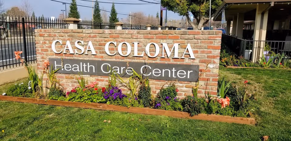 Brick sign reading "CASA COLOMA Health Care Center" on a grassy lawn with flowerbeds in front of the facility.