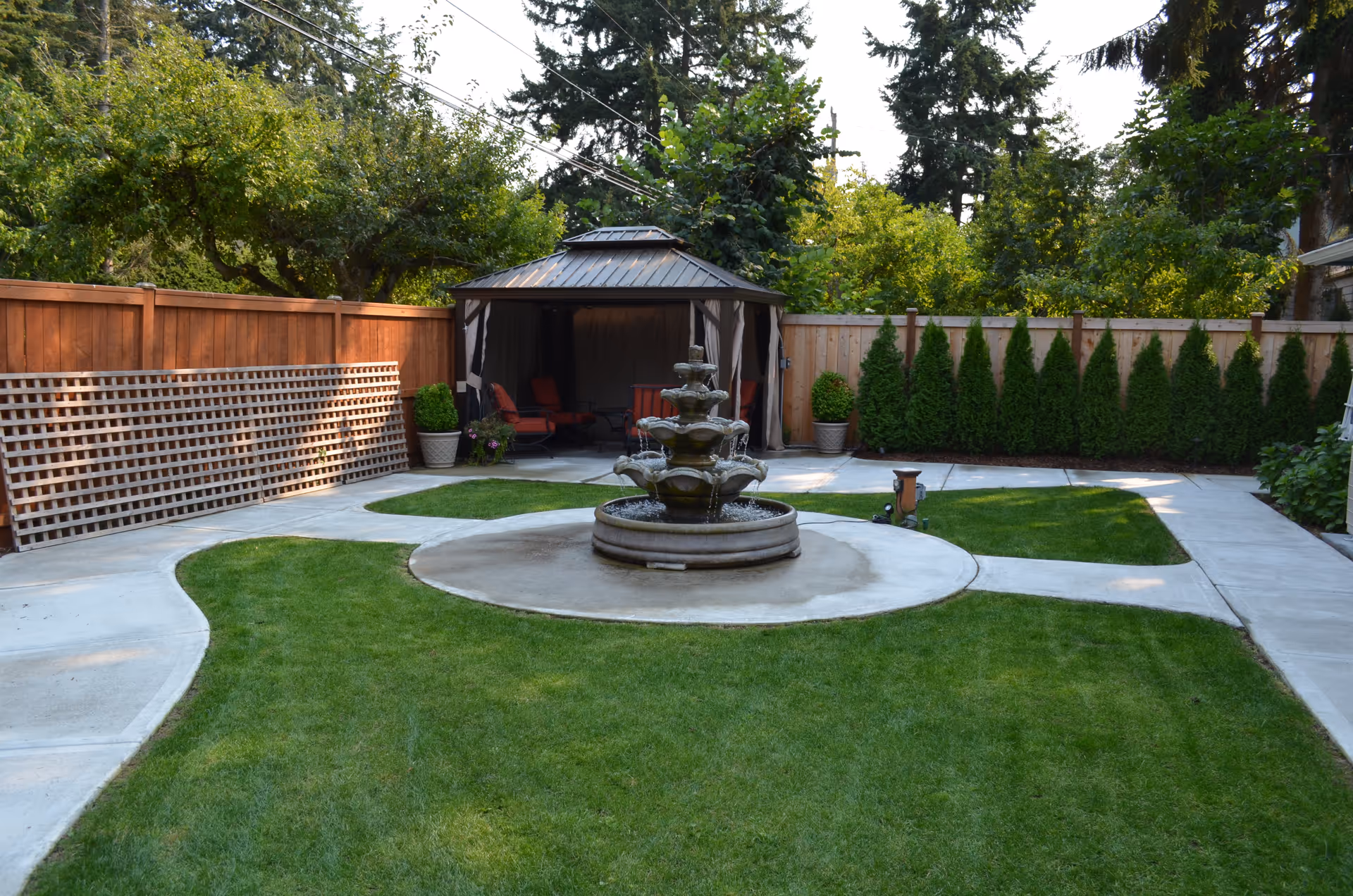 A well-maintained outdoor garden area with a central multi-tiered water fountain surrounded by green grass and concrete pathways. There is a wooden fence in the background with tall evergreen shrubs and a gazebo with seating inside.