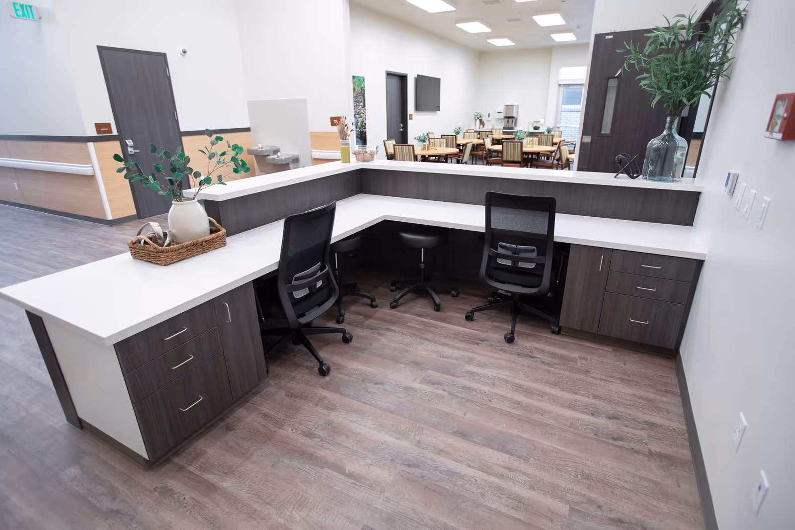 A modern reception desk area with two black office chairs and a small black stool behind the counter. The desk has a white countertop and dark wood cabinetry. There are decorative plants on the desk, and in the background, there is a dining area with tables and chairs, as well as doors and a water fountain on the wall.
