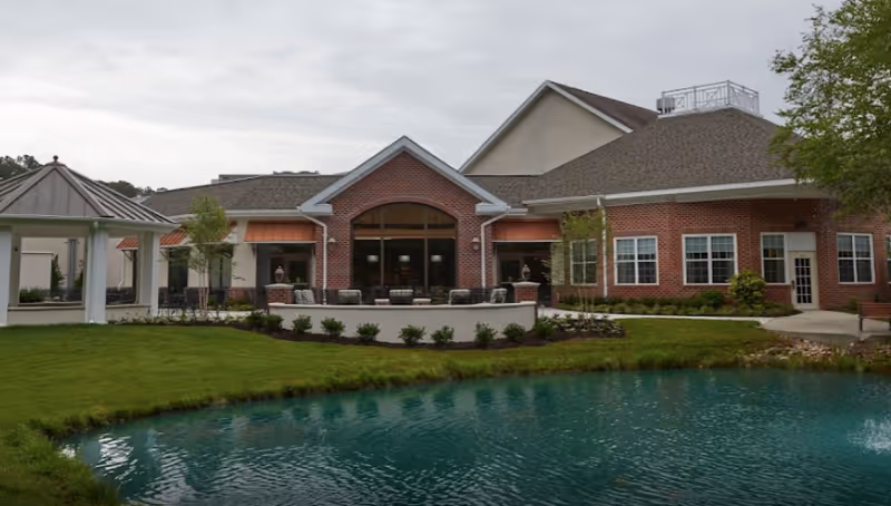 Brick-front senior living facility with a patio seating area and a pond in the foreground.