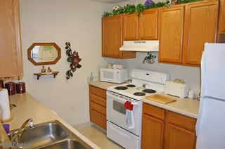 A compact kitchen featuring wooden cabinets, a white electric stove with an oven, a microwave, a white refrigerator, and a stainless steel sink. The countertop holds a cutting board, a toaster, and some decorative items. There is a small wall shelf with decorative pieces and a mirror on the wall.