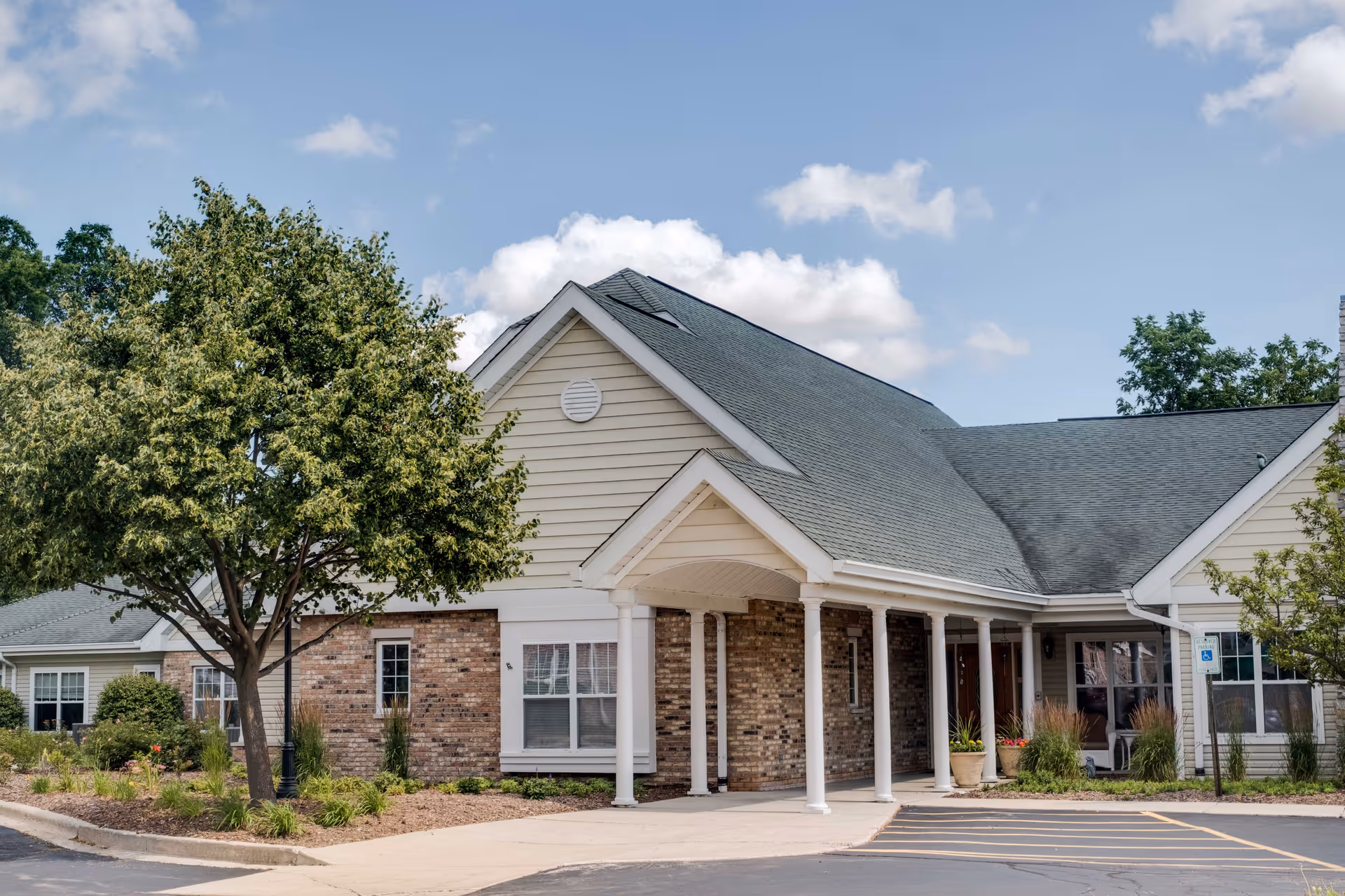 Exterior view of a single-story assisted living facility building with a covered entrance supported by white columns, surrounded by landscaping including trees and shrubs under a partly cloudy sky.