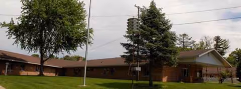 Single-story brick facility with green lawn, trees, and a flagpole in front.