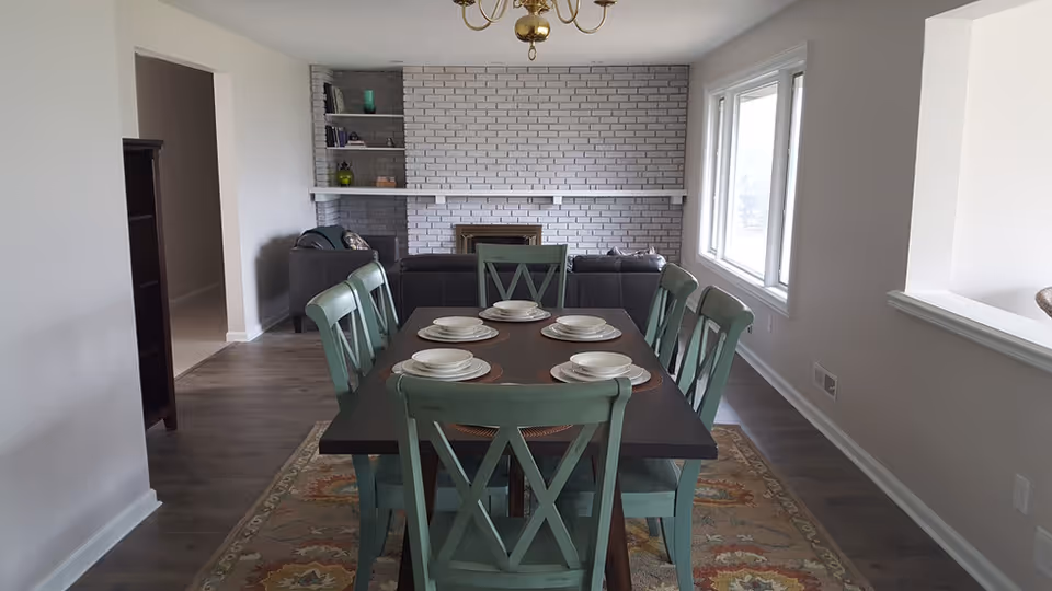 Dining area with a dark wooden table set with six place settings including plates and bowls. Six green wooden chairs surround the table. Behind the dining area is a living room with a dark sofa facing a white painted brick fireplace with built-in shelves on the left. Large windows on the right side let in natural light. The floor is wood with a patterned area rug under the dining table.