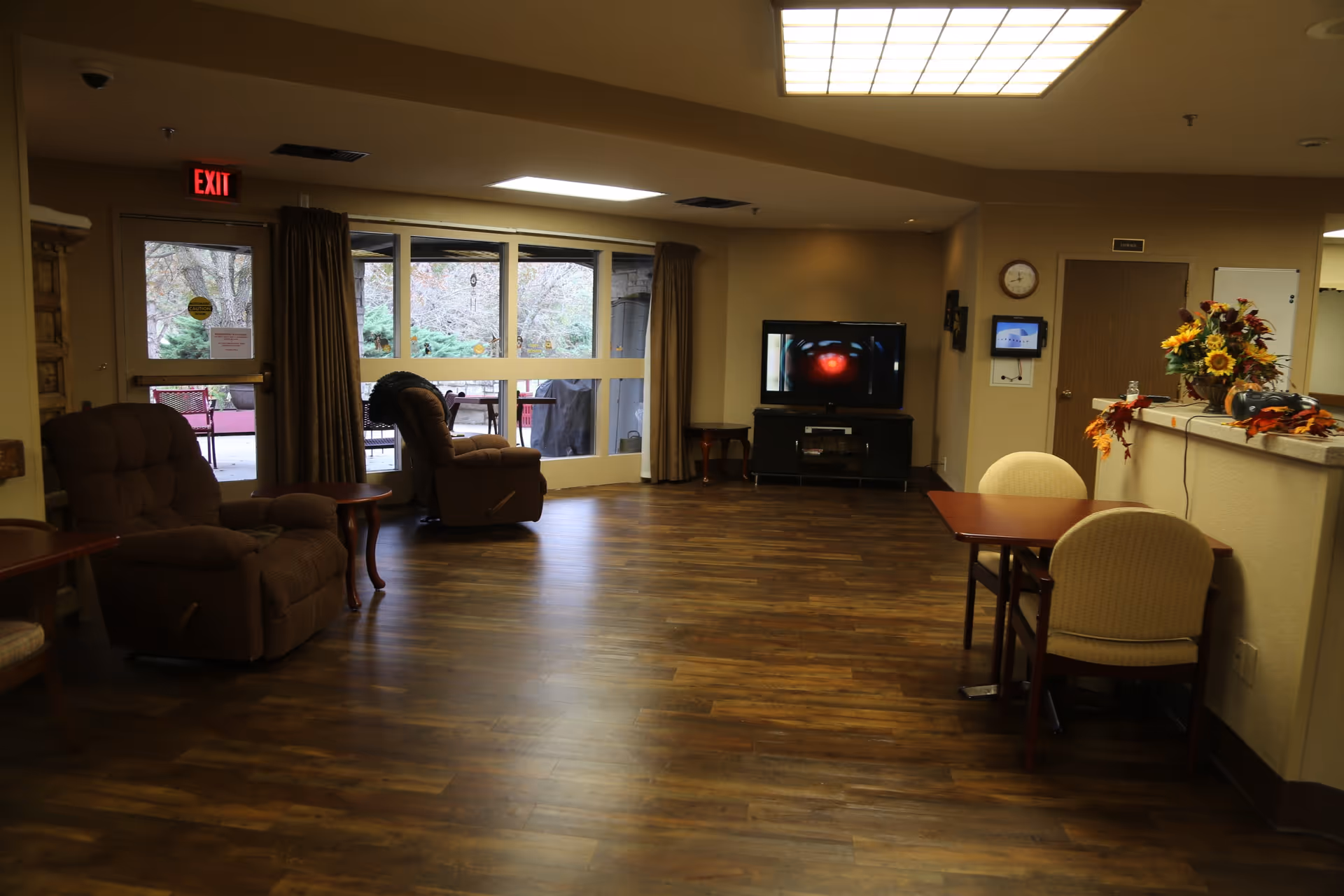 A cozy common area in a senior living facility with wooden flooring, two brown recliner chairs near a glass door leading outside, a flat-screen TV on a stand against the wall, a small table with two beige chairs, and a counter decorated with a vase of sunflowers and autumn leaves.