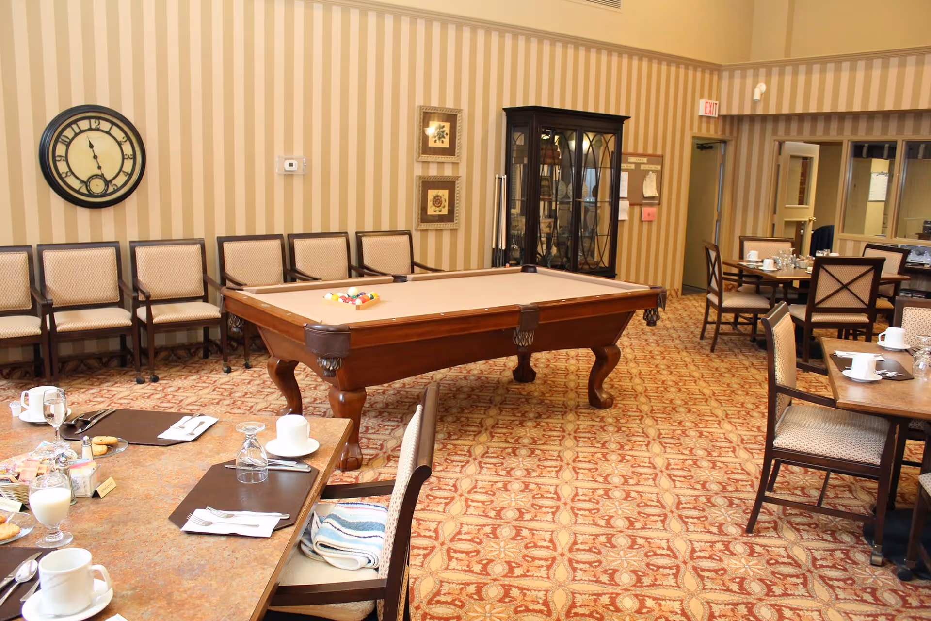 A room with a pool table in the center surrounded by chairs along the wall and dining tables set with cups, glasses, and plates. The room has patterned carpet and striped wallpaper with a large clock on the wall and a glass cabinet in the corner.