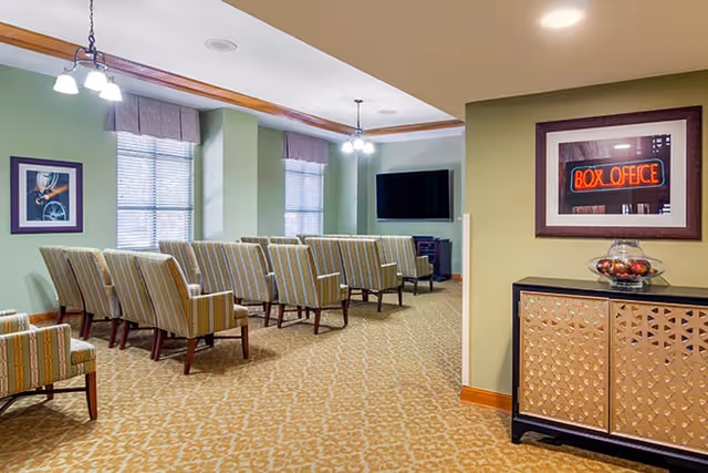 A small theater-style room with several rows of striped upholstered chairs facing a large flat-screen TV mounted on the wall. The room has green walls, patterned carpet, two windows with blinds and valances, and ceiling light fixtures. On the right side, there is a cabinet with a decorative bowl on top and a framed picture of a neon 'BOX OFFICE' sign above it.