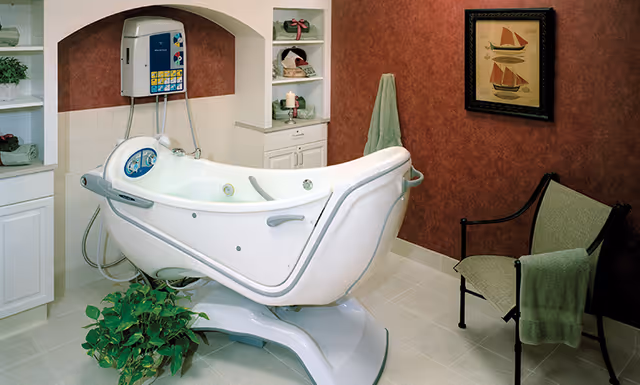 A white assisted bathing tub in a private bathroom with shelving, a chair, towels, a potted plant and a framed sailboat picture on the wall.