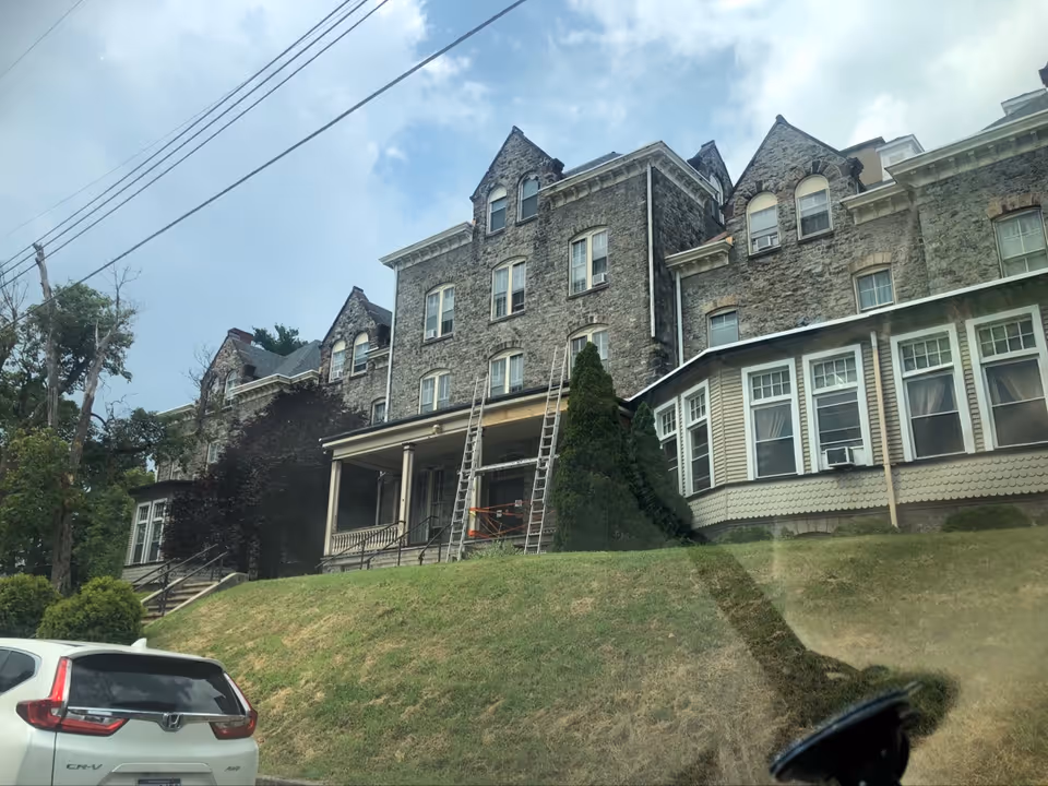 A large, multi-story stone building with a steep roof and multiple windows, identified as Stabon Manor Personal Care Home. The building has a porch with columns and ladders leaning against it, suggesting maintenance work. There is a grassy hill in front of the building and a white Honda CR-V parked at the bottom left. The sky is partly cloudy.