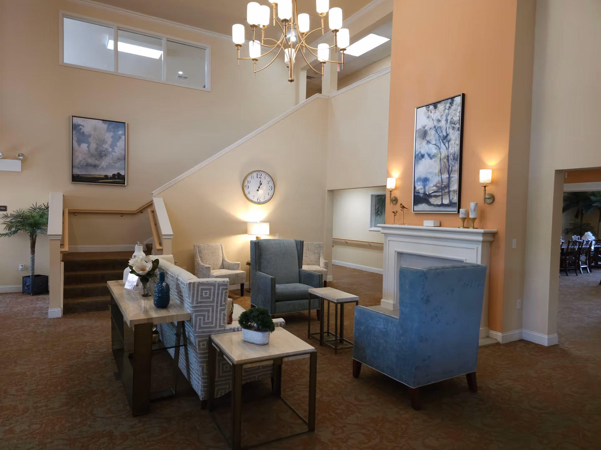 A spacious and well-lit living room area with high ceilings, featuring a chandelier, a fireplace with wall sconces and a painting above it, several upholstered chairs and sofas arranged around small tables, a clock on the wall, and a staircase leading to an upper level.