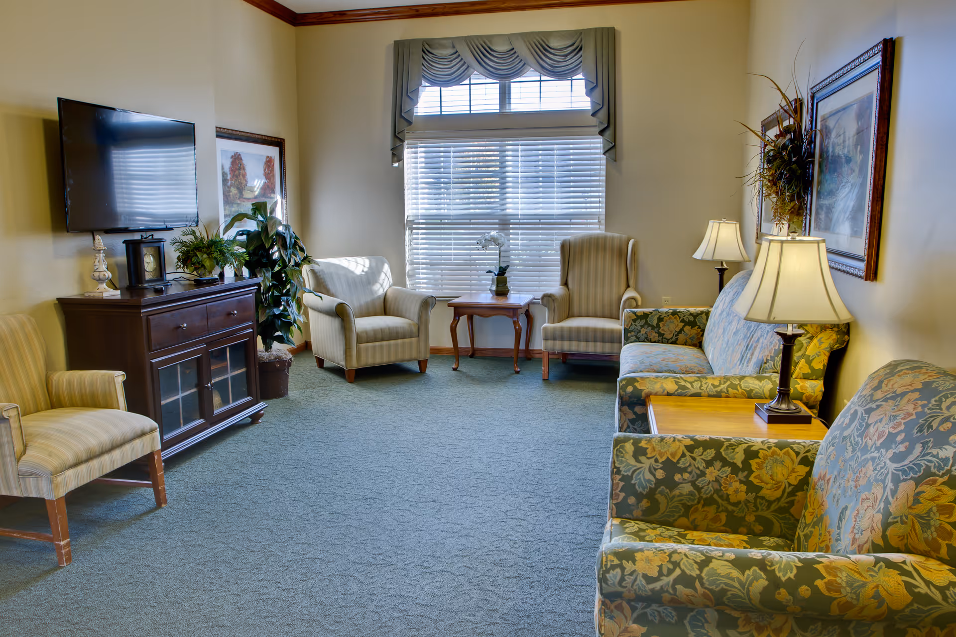A cozy living room with floral patterned sofas, striped armchairs, a wooden cabinet with a TV mounted above it, a small wooden table with a potted plant, and framed artwork on the walls. The room has a large window with blinds and a decorative valance, and two table lamps providing warm lighting.