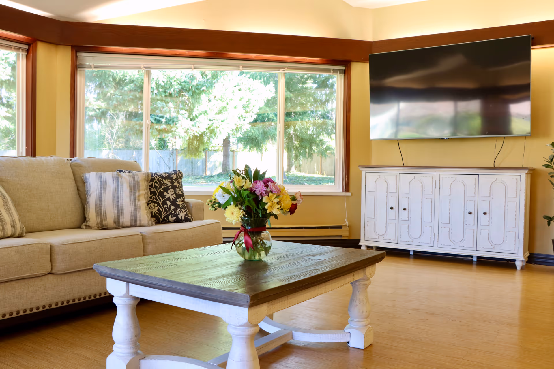 A bright living room with a beige sofa adorned with patterned pillows, a wooden coffee table with a vase of colorful flowers, a large window showing green trees outside, and a flat-screen TV mounted on a yellow wall above a white cabinet.