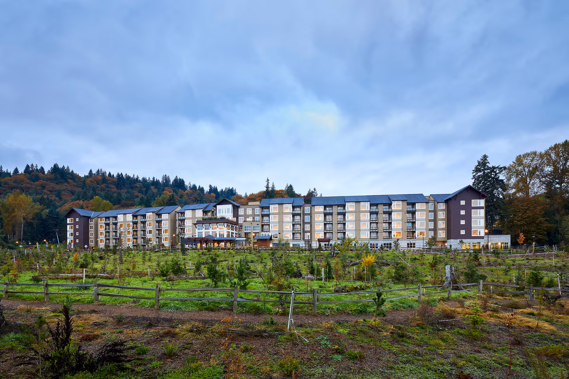 Wide exterior view of a multi-story senior living facility named Revel Issaquah, surrounded by a green landscaped area with young trees and a wooden fence, set against a backdrop of forested hills under a cloudy sky.