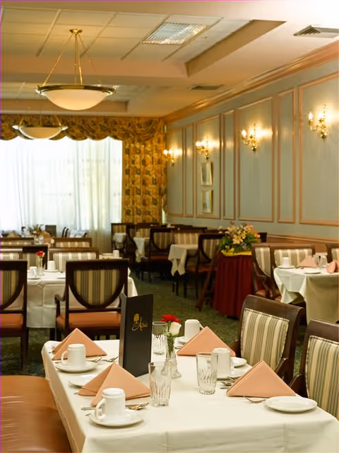 A well-lit dining room with tables covered in white tablecloths, set with cups, plates, glasses, and folded pink napkins. The room features striped upholstered chairs, wall sconces, framed pictures, and large windows with patterned curtains allowing natural light to enter.