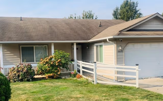 Front of a single-story house with a garage, a wheelchair ramp leading to a small covered porch, and a lawn with shrubs.